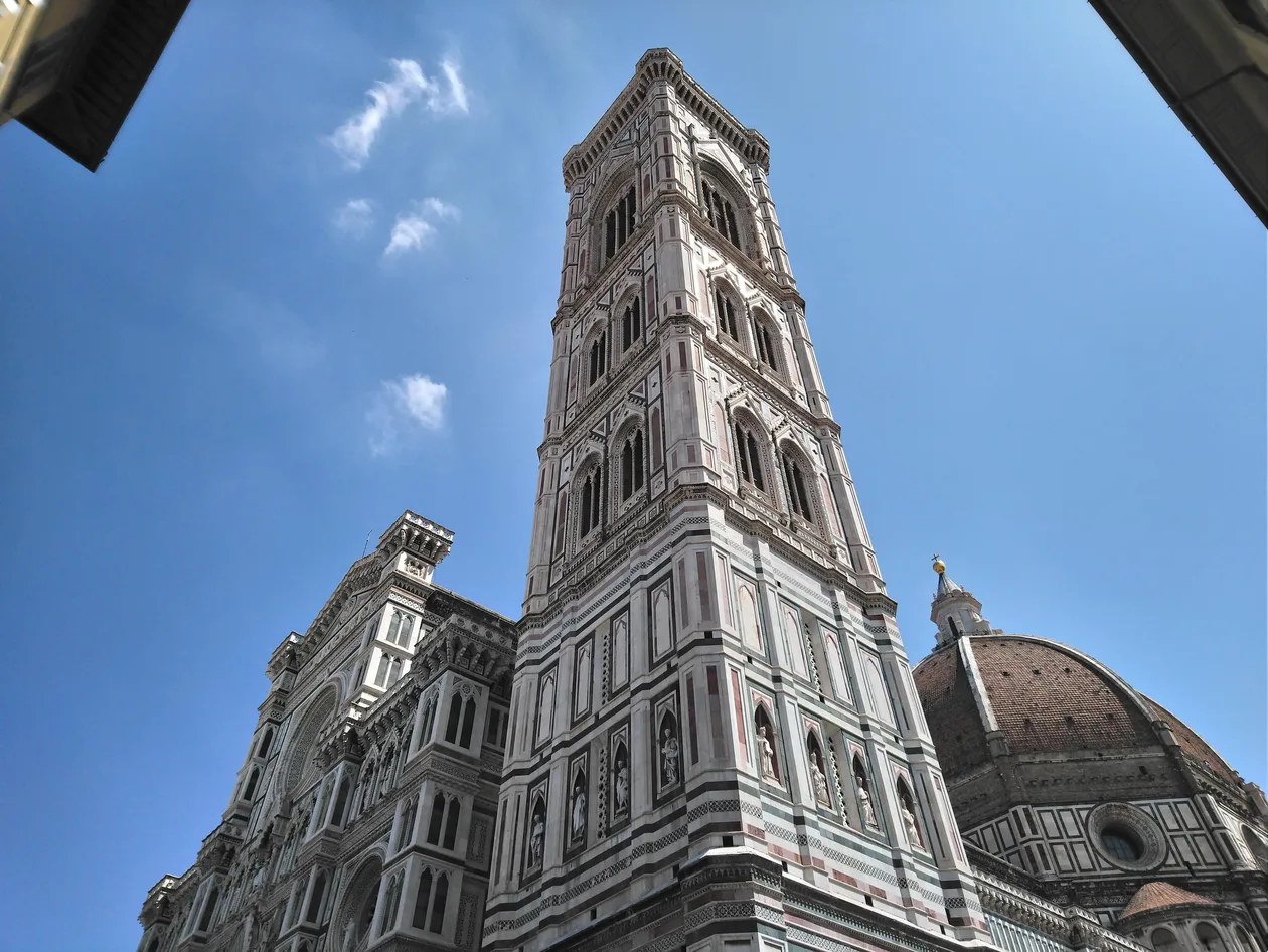 Giotto's Bell Tower seen from below against a blue sky with a few clouds, the cathedral facade at its foot on the left and Brunelleschi's dome on the right, framed by dark rooftops in the top corners