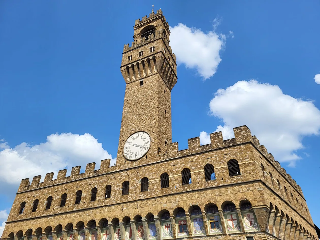The Arnolfo tower of Palazzo Vecchio rising above the battlements and the clock face, seen from below against a blue sky with white clouds