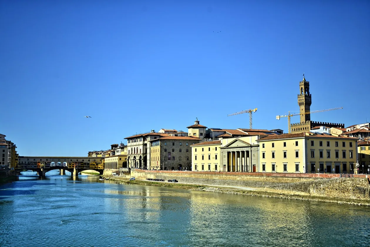 The Arno in Florence under a clear sky, the Uffizi and the riverside buildings on the right bank with Palazzo Vecchio's tower behind, and the Ponte Vecchio in the distance