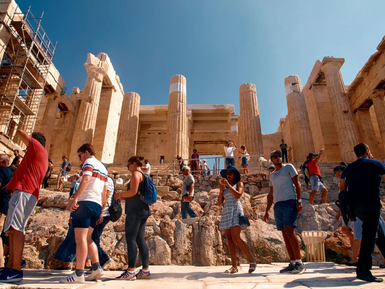 Visitors on the rocky path below the Propylaea gateway of the Acropolis, with scaffolding on the left columns, under a clear blue sky