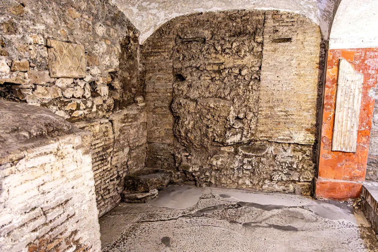 A vaulted underground Roman room with rough brick walls and a fragment of red-framed wall painting