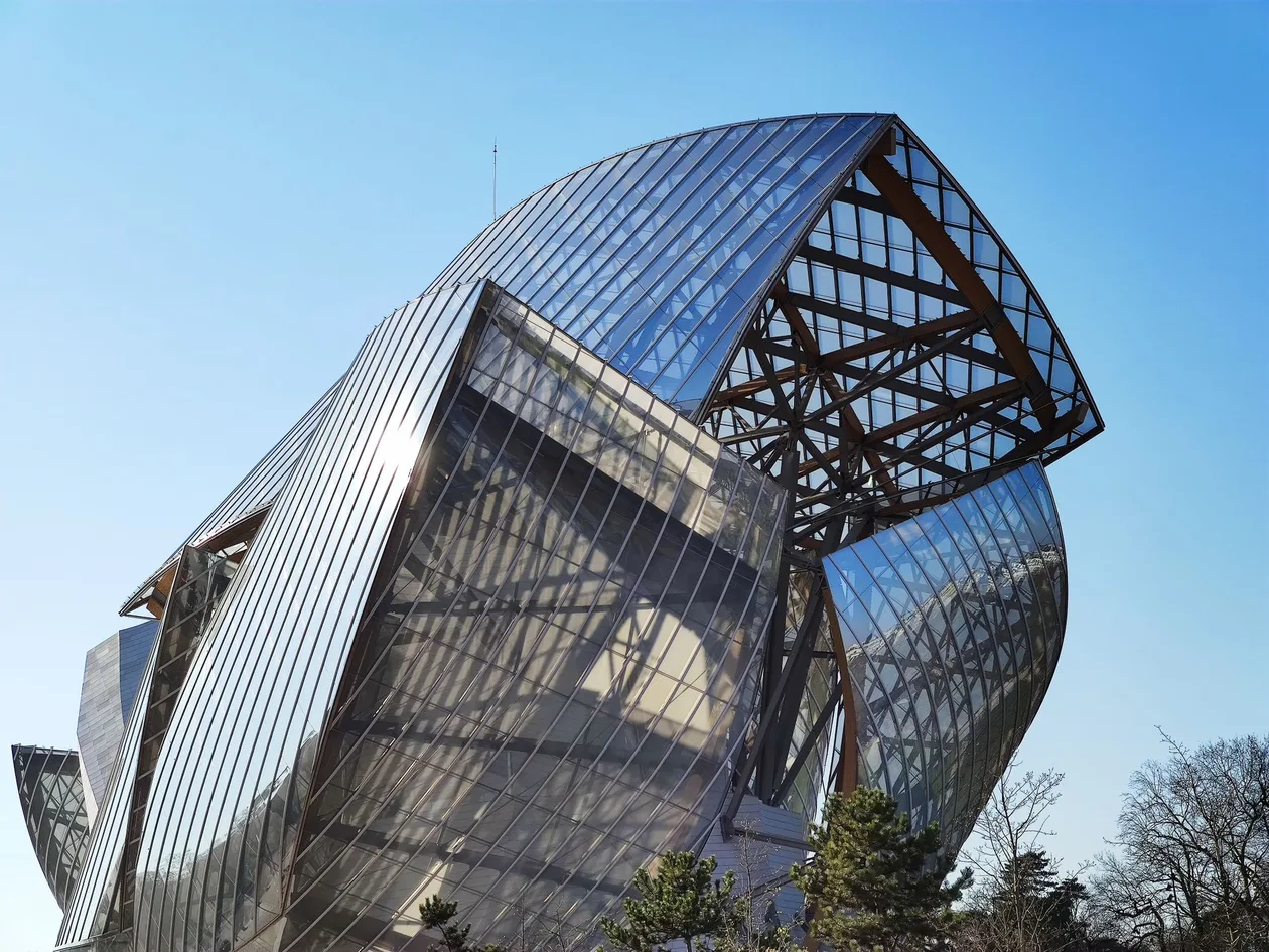 Curved glass sails and timber beams of Frank Gehry's Fondation Louis Vuitton against a clear blue sky, pine trees in the foreground