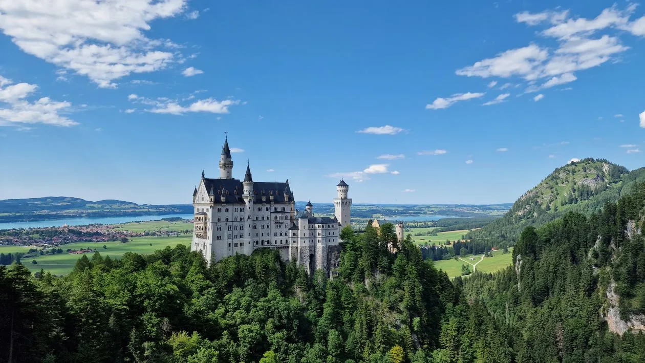 Neuschwanstein Castle on its wooded ridge in summer, seen from the Marienbrücke side, with the Forggensee lake and Bavarian plain behind