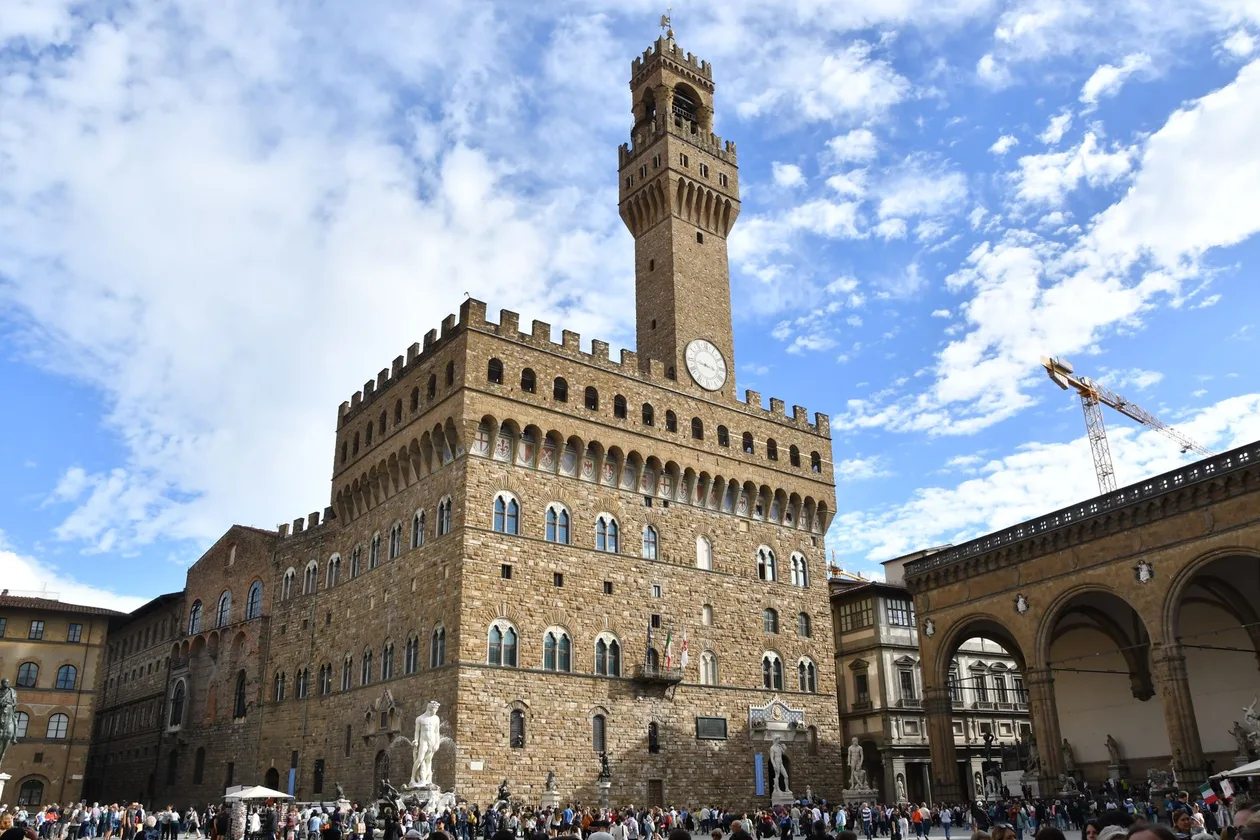 Palazzo Vecchio seen from Piazza della Signoria under a blue sky, the Arnolfo tower rising above the battlements and the square below full of visitors around the Neptune fountain and the Loggia dei Lanzi
