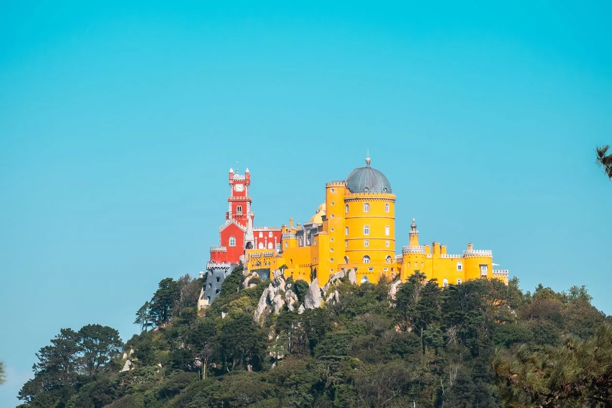 Pena Palace on its wooded hilltop in Sintra, the red clock tower and the yellow domed tower rising above granite outcrops against a clear blue sky