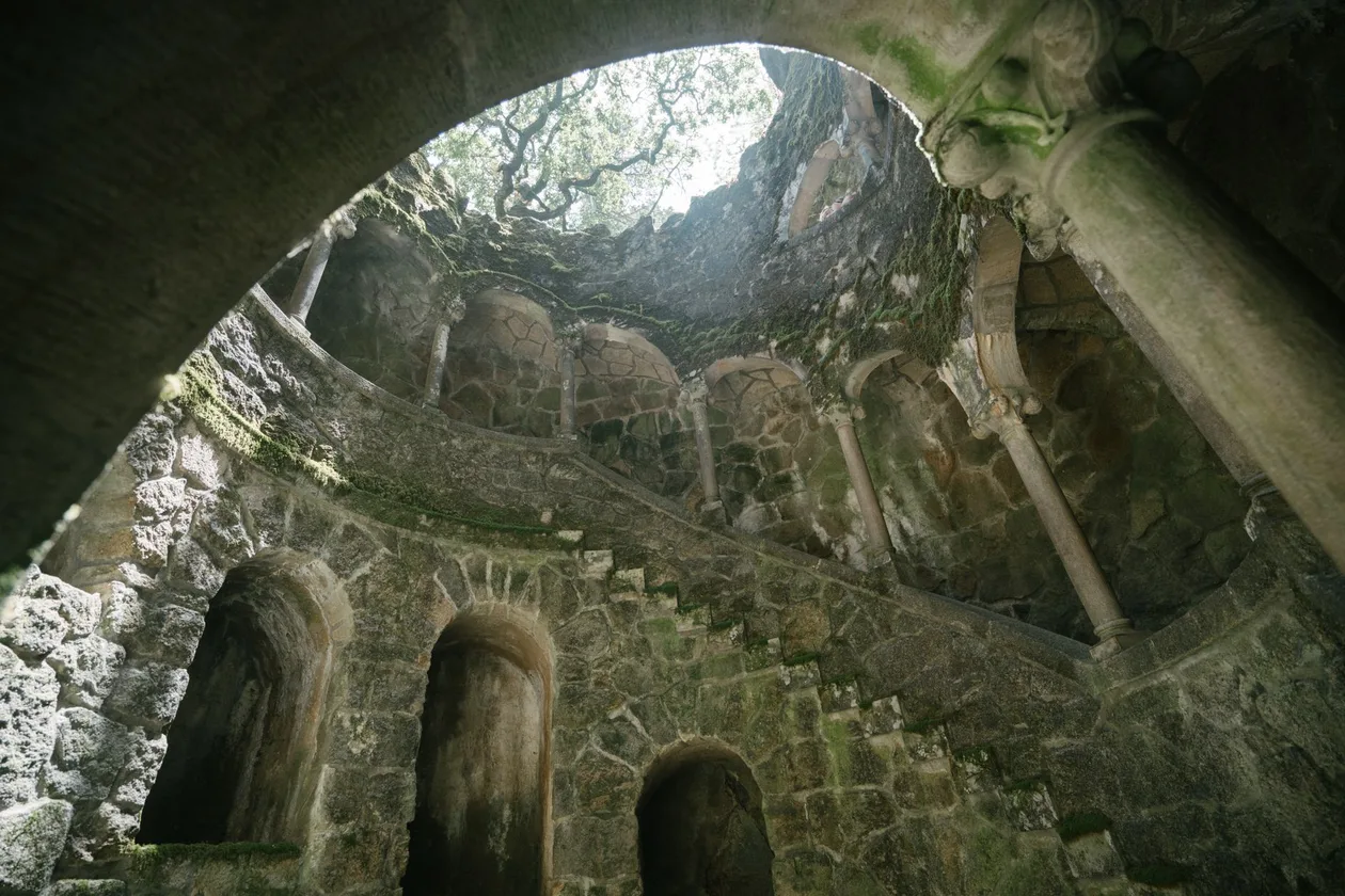 Looking up from inside the Initiation Well at Quinta da Regaleira, mossy stone galleries with columns spiralling up to a circle of trees and daylight